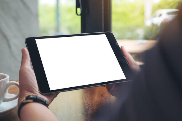 Falešný obraz ženských rukou držících černý tablet PC s bílou prázdnou obrazovkou a šálkem kávy na dřevěném stole v kavárně - Fotografie, Obrázek