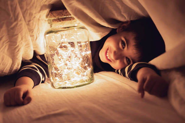 Criança joga sob a folha com um pote de luzes quentes Conceptual de estilo de vida, jogo infantil em casa - Foto, Imagem