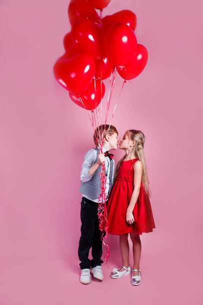 Valentines day. Little boy kissing beautiful little girl on pink background. Children holding heart balloons - Фото, изображение