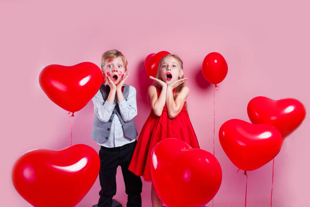 Funny little boy with red kisses on the skin and girl in red balloons in the shape of a heart on pink background - Photo, image