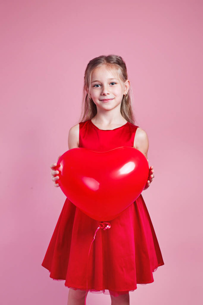 Valentines day. Little girl in red dress with heart shape air balloon on pink studio background - Photo, Image