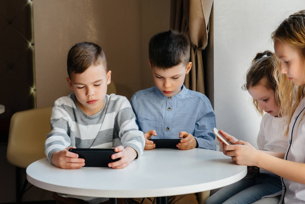 Children sit at a table in a cafe and play mobile phones together. Modern entertainment - Foto, Imagem