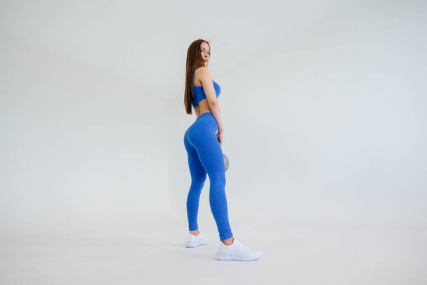 Sexy young girl performs exercises with a weight in a blue tracksuit on a white background. Fitness, healthy lifestyle - Fotografie, Obrázek