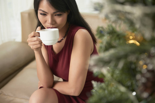 Gyönyörű fiatal ázsiai nő ül kanapén közel karácsonyfa és ivás csésze kamilla tea - Fotó, kép