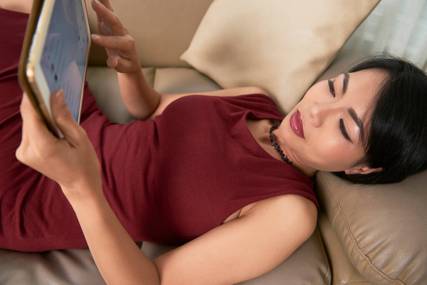 Приваблива молода в'єтнамська жінка лежить на дивані і читає повідомлення друзів у соціальних мережах. - Фото, зображення