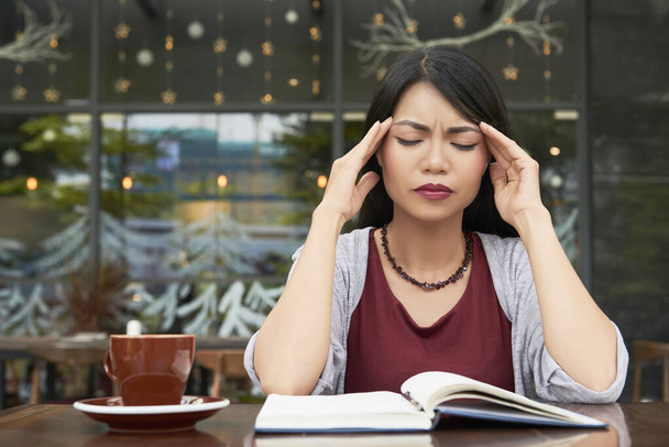 Портрет красивой молодой азиатки, страдающей от сильной головной боли при попытке работать в кафе - Фото, изображение