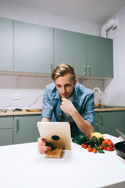 Young caucasian man use tablet computer in the modern kitchen, preparing salad, read recipe. - Fotoğraf, Görsel