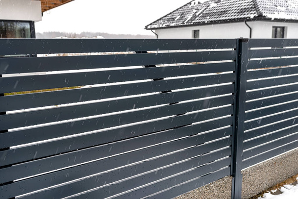 Valla de panel antracita moderna con un conector visible de los cimientos de la valla, llueve en invierno, en el fondo los techos de la casa con nieve. - Foto, Imagen