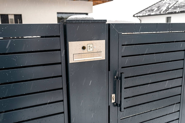Современный видеотелефон с почтовым ящиком и беспроводным кард-ридером, смонтированным в панельном заборе антрацитного цвета, видимой калиткой, зимой идет дождь, на заднем плане крыши дома со снегом. - Фото, изображение