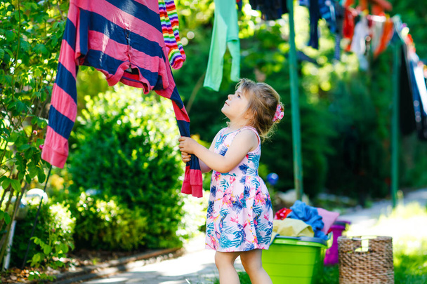 Toddler girl hanging clothes, fresh clean dresses and trousers for drying in garden, outdoors after making laundry. Happy child helping in household. Family working together, kid learning to help. - Photo, Image