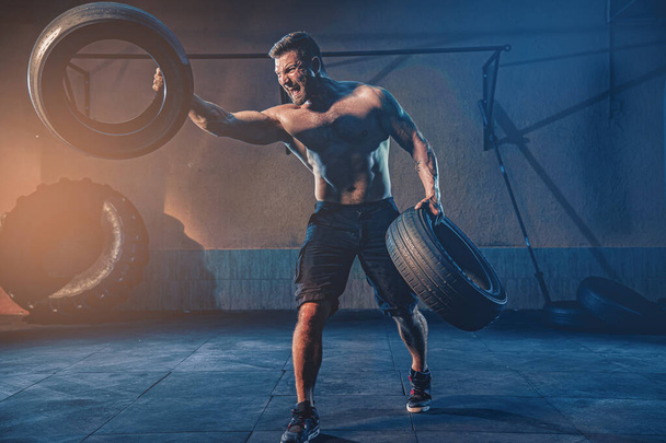 Bodybuilding training, bearded strong sportsman with muscular body lifting heavy wheel in gym - Fotó, kép
