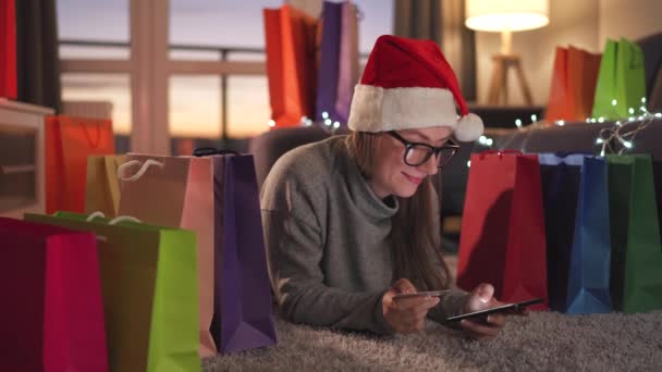 Mujer feliz con gafas con un sombrero de Santa Claus está acostado en la alfombra y hace una compra en línea con una tarjeta de crédito y un teléfono inteligente. Bolsas de compras alrededor. - Metraje, vídeo