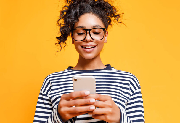 Young ethnic female in glasses smiling and browsing social media on smartphone against yellow background - Фото, зображення