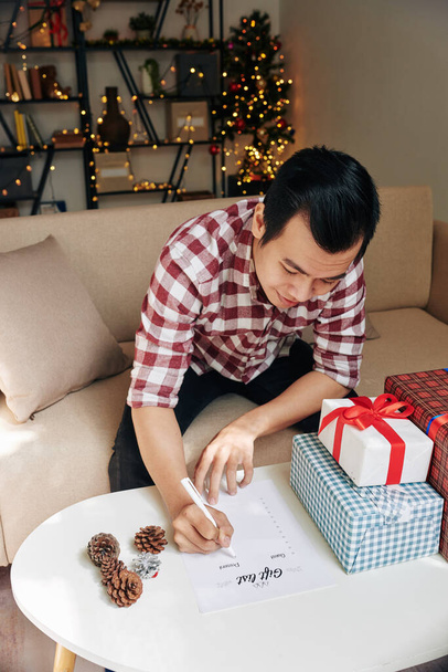 Sorrindo jovem asiático homem preenchendo lista de presentes e embrulhando presentes de Natal para parentes e amigos - Foto, Imagem
