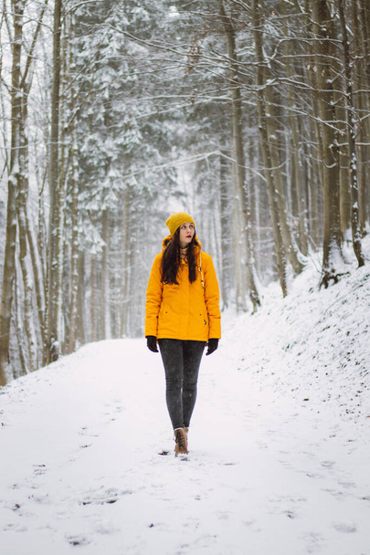 Μελαχρινή γυναίκα περπατά μέσα από ένα χιονισμένο τοπίο και απολαμβάνει την ομορφιά του. Κορίτσι με κίτρινο μπουφάν και τακούνια. Πορτρέτο της αυτοπεποίθηση αθλητριών σε αντίκες λευκό τόνο. 20-24 ετών. - Φωτογραφία, εικόνα