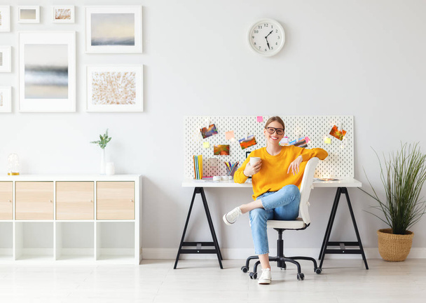 Smiling female entrepreneur sitting on chair near table in cozy home workspace and enjoying break with cup of coffee - Photo, image