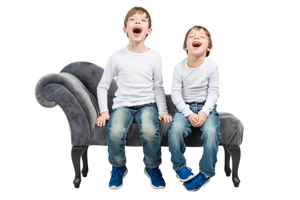 Portrait of two young boys with their mouth open, sitting on a couch, isolated on white background - Fotó, kép