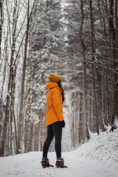 mujer de entre 20 y 24 años parada en medio de una carretera nevada y nevando a su alrededor. Retrato en blanco antiguo y tono champán de vela. Chica en una chaqueta de invierno amarillo y vaqueros negros. - Foto, imagen