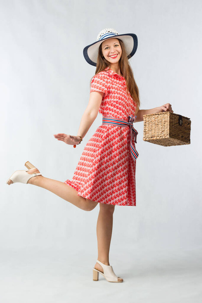 Young cheerful woman in red dress posing with picnic basket in studio on white background - 写真・画像