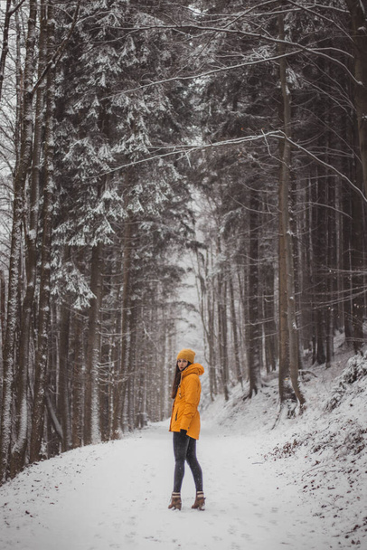 Νεαρή κοπέλα με κίτρινο χειμωνιάτικο μπουφάν και με ένα ακατανόητο χαμόγελο κοιτάζει το σύντροφό της. Γυναίκα ηλικίας 20-24 ετών στέκεται στη μέση του χιονισμένου δρόμου και χιονίζει γύρω της. Σετ Σαμπάνια. - Φωτογραφία, εικόνα