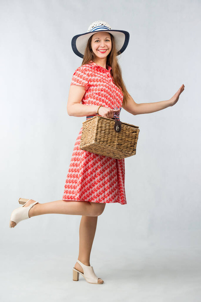 Young cheerful woman in red dress posing with picnic basket in studio on white background - Fotó, kép