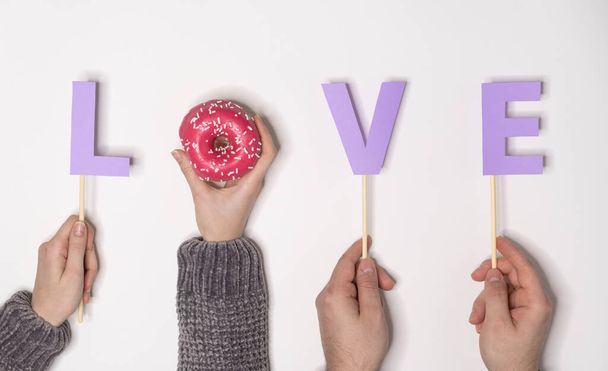 Top view of the word LOVE made of purple letters and a donut with pink filling on a white background. Valentine's day card. - Фото, изображение