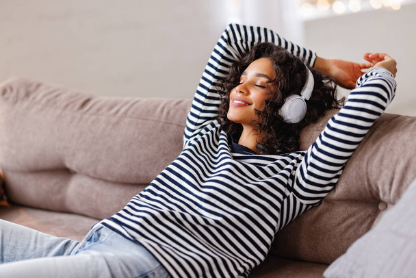 Mujer étnica feliz con los ojos cerrados sonriendo y escuchando música favorita en los auriculares mientras se relaja en el cómodo sofá en casa - Foto, imagen