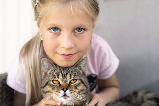 Χαριτωμένο αξιολάτρευτο καυκάσιο ξανθό γλυκό κοριτσάκι οκτώ ετών απολαμβάνουν κάθεται σε πολυθρόνα εξωτερική εκμετάλλευση και αγκαλιάζει μικρή γάτα υπνάκο στα χέρια. Έννοια φιλίας για γάτες και παιδιά. Φροντίδα και αγάπη των ζώων - Φωτογραφία, εικόνα