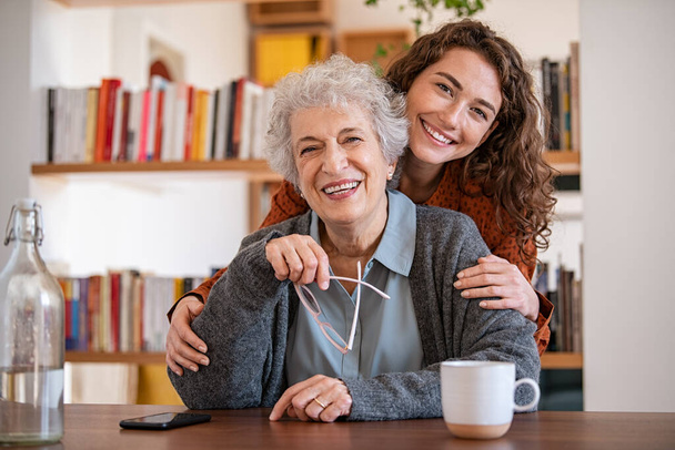 Cheerful young woman embracing senior mother at home and looking at camera. Portrait of happy adult granddaughter and grandmother embracing and smiling together. Portrait of lovely young woman hugging from behind elderly granndma. - Foto, Bild