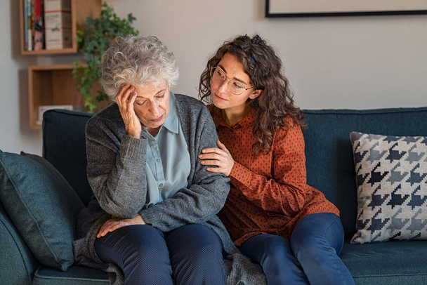 Caring daughter comforting frustrated unhappy senior woman. Loving adult granddaughter talking to sad depressed old grandmother holding hand and comforting her. Upset widowed woman with headache consoled by her daughter. - 写真・画像