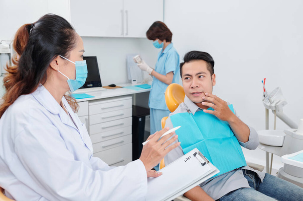 Dentista discutiendo dolor de muelas con el paciente cuando la enfermera prepara herramientas en segundo plano - Foto, imagen