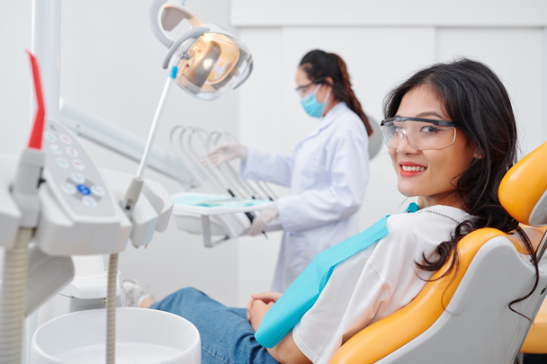 Pretty smiling young Vietnamese woman in goggles sitting in dental chair and looking at camera - Photo, Image