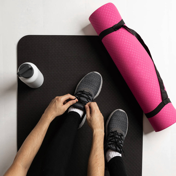 A top view of a man tying shoe laces on a black fitness mat with a thermos water bottle - Фото, изображение