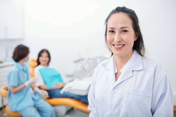 Neşeli deneyimli Asyalı ortodontist kameraya gülümsüyor, asistanı genç bir hastayla konuşuyor. - Fotoğraf, Görsel