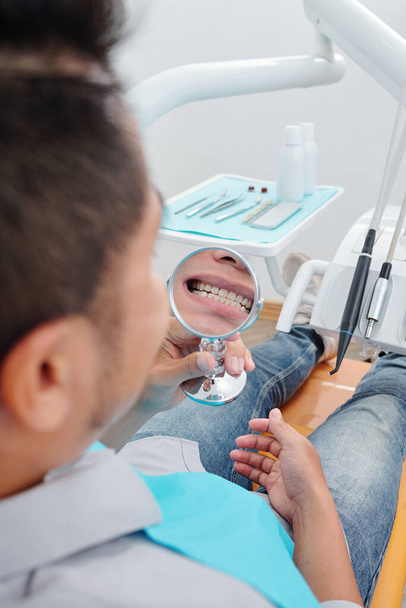 Patient looking in mirror at his treated teeth when sitting in dental chair - Foto, Imagen