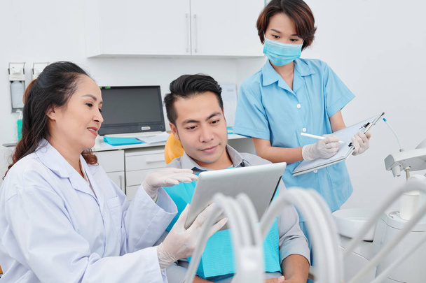 Dentiste et patient discutant de la radiographie de la mâchoire sur tablette numérique lorsque l'assistant remplit les informations dans le document - Photo, image