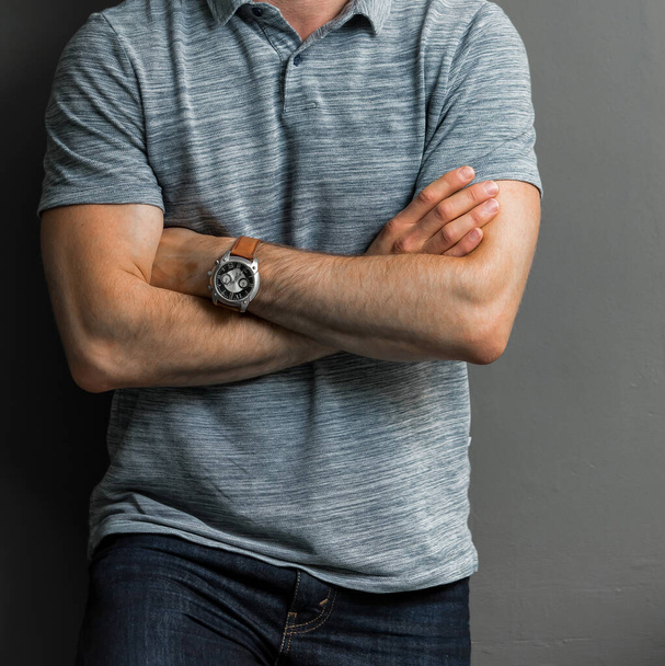 A young man in a gray shirt wearing a watch with his arms crosse leaning on a wall - Foto, Imagem