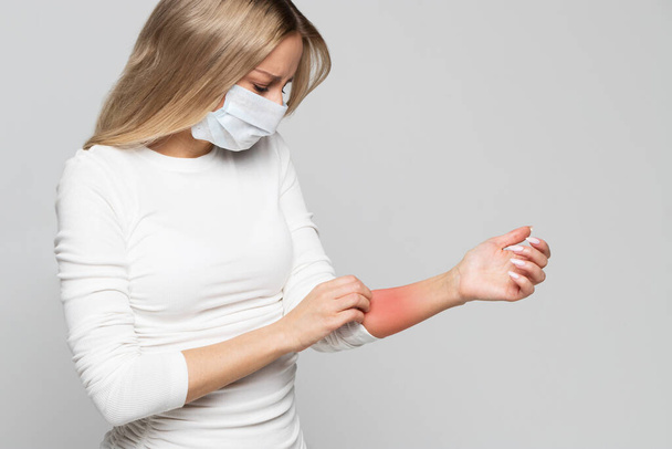 Studioporträt einer kranken jungen Frau in Schutzmaske, die sich den Arm kratzt, isoliert auf grauem Hintergrund. Symptome einer Tierallergie, juckende Haut - Foto, Bild