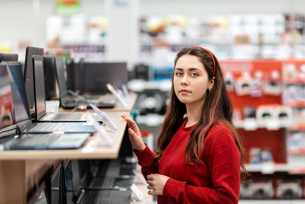 Młoda brunetka w czerwonej bluzie wybiera laptopa w sklepie. Koncepcja zakupu dóbr cyfrowych. - Zdjęcie, obraz