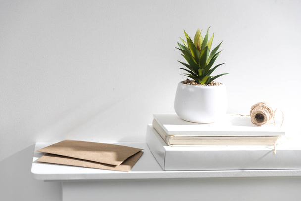 An artificial succulent plant in a white ceramic vase stands on boxes and notebooks. A skein of craft twine. Two brown envelopes. - Foto, imagen