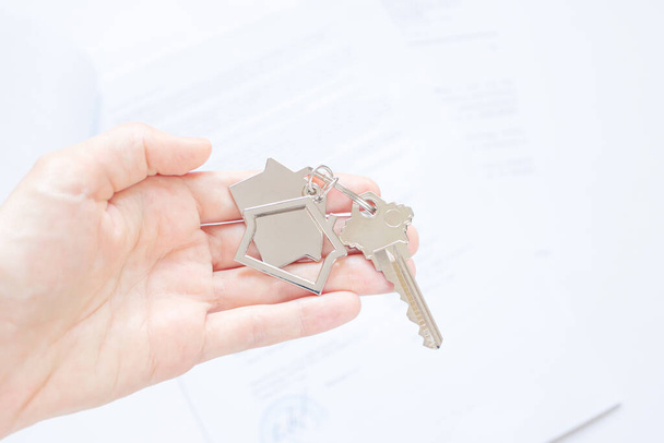Close-up of a key with a house-shaped keychain in the palm of the hand, documents in the background. The concept of buying and insuring real estate, apartments, mortgages. Light colors, copy space. - Photo, Image