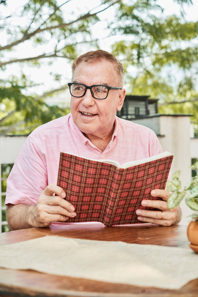 Portrait of cheerful senior man in glasses sitting outdoors at table with textbook in hands, learning every age concept - Photo, Image