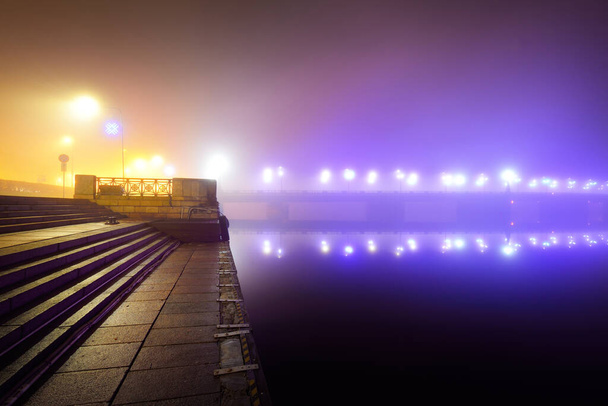 Iluminado puente de piedra vacío en una niebla por la noche. Primer plano de las linternas. Reflejos de simetría en el agua, espejo natural. Río Daugava, Riga, Letonia. Imagen conceptual, colores neón - Foto, Imagen