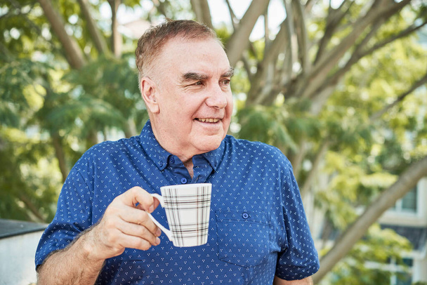 Портрет усміхненого старшого чоловіка, що стоїть на відкритому повітрі з чашкою кави і дивиться в сторону
 - Фото, зображення