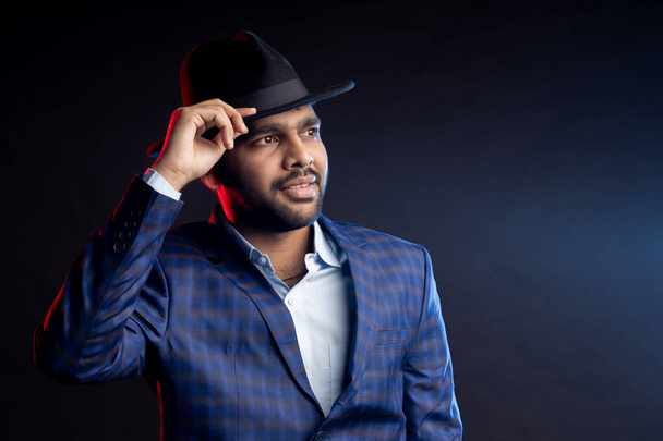 Studio portrait of young handsome confident unshaven Indian man, businessman wearing shirt, checked suit, looking at camera, touching hat, smiling, standing isolated on dark background. - Fotografie, Obrázek