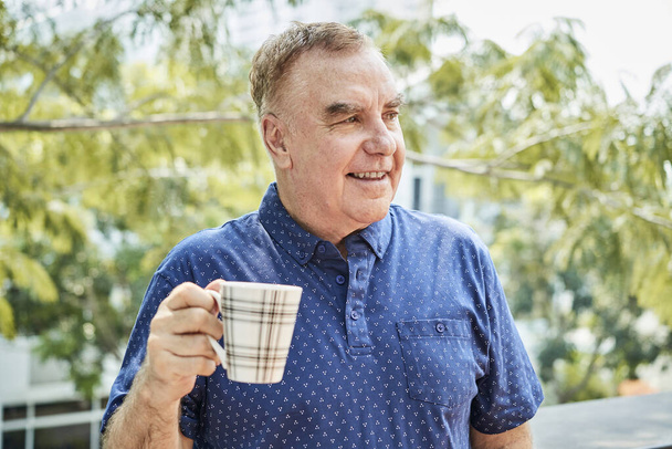 Portrét pozitivního staršího muže stojícího venku s šálkem kávy a dívajícího se stranou - Fotografie, Obrázek
