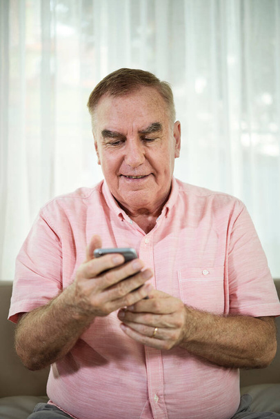 Portrait of smiling senior man reading text messages in smartphone when sitting on sofa in living room - Фото, зображення