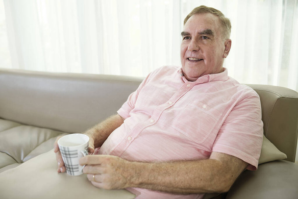 Retrato de un hombre mayor positivo relajándose en el sofá con una taza de café o té y viendo la televisión - Foto, imagen