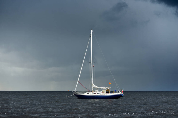 Yate amañado balandra azul anclado frente a la costa de Inglaterra bajo el cielo oscuro después de una tormenta eléctrica. Navegando con mal tiempo. Paisaje marino dramático. Rayos de sol a través de las nubes - Foto, Imagen