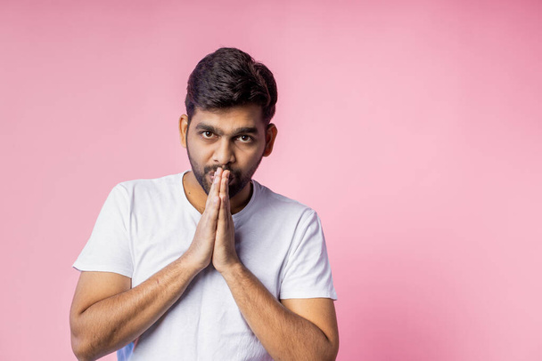 Young religious indian unshaven man wearing white t shirt, keeping palms together, looking down, praying, hoping for best, isolated over in pink background. Belief concept. - Fotografie, Obrázek